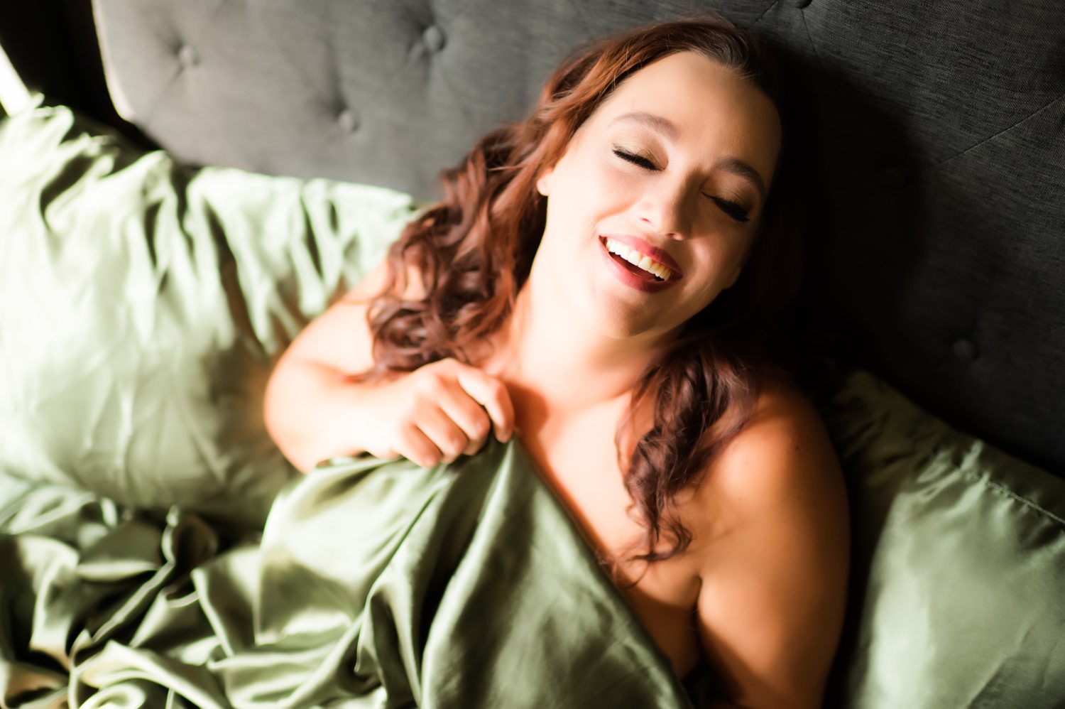Sexy photo of woman smiling sitting in bed holding a green sheet to her chest.