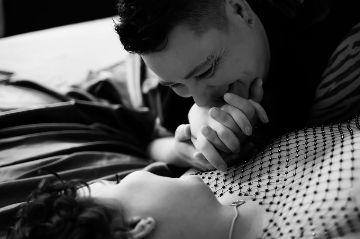 Black and white image of a person kissing the hands of her partner while lovingly looking over them while they are both lying down on their bed