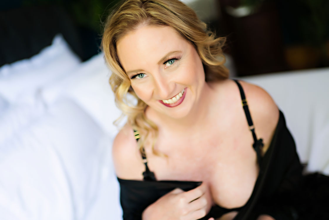 Woman wearing a black lingerie and black cover up while sitting in a white bed for a boudoir shoot