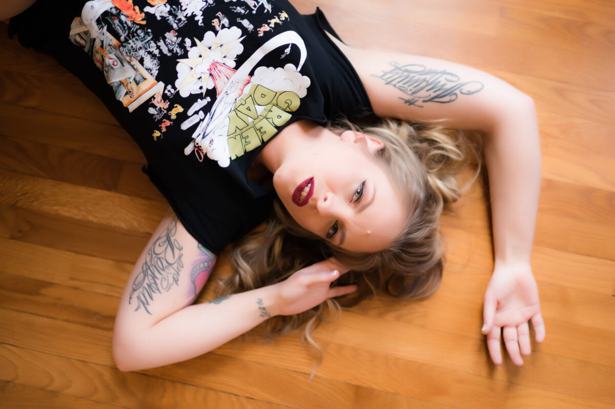A woman with tattoo on both her arms laying down on the floor, wearing a black shirt and posing for a boudoir shot