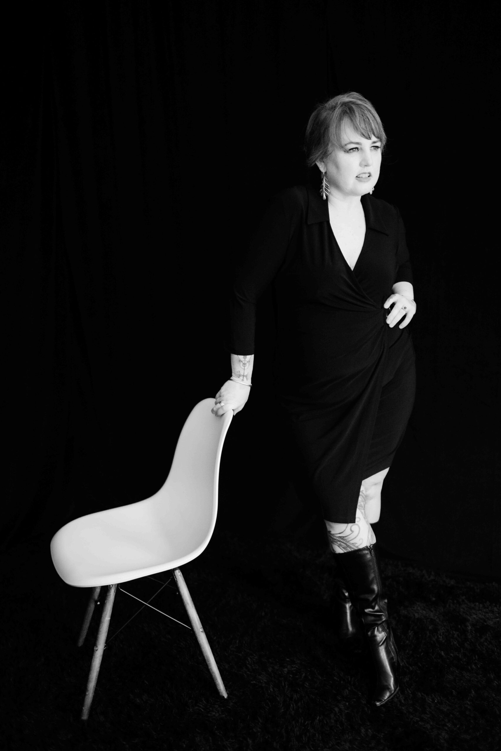 Black and white photo of a woman in black dress. She's standing beside a chair and her hand is resting on the chair