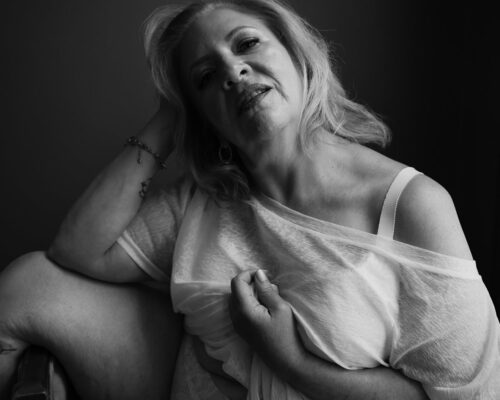 Sexy black-and-white portrait of woman in her 50s sitting with her leg over an antique chair.
